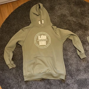 Olivgrön hoodie från Björn Borg - Säljer en olivgrön hoodie från Björn Borg med vit logga och text på bröstet. Tröjan har huva med dragsko och en stor magficka framtill. Perfekt för en avslappnad stil.
