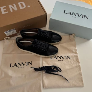 Svarta sneakers från Lanvin med lackad tå - Säljer ett par svarta sneakers från Lanvin med snygg lackad tå och ovandel i mocka. Skorna har svarta snören, vita sulor och klassisk rund tå. Perfekta för dig som gillar stilrena och exklusiva sneakers.