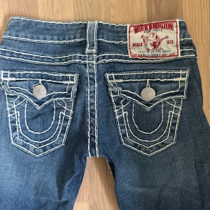 W23 True Religion jeans vintage y2k m vita sömmar - Vintage jeans från True Religion med smala ben, ej helt skinny men inte vida. Kan klippas upp nedtill för bootcut eller göra shorts/kjol av. Tydliga vita kontrastsömmar och stora bakfickor med dekorativa knappar. Svåra att få tag på! Klassisk femficksmodell och rakt snitt. Märkespatch bak i rött och vitt. Perfekta för dig som gillar statement-jeans. Storlek w23 (23 tum i midjan) motsvarar ungefär strl 32/xs. Innerbenslängd: 77 cm. Ytterben: 97 cm