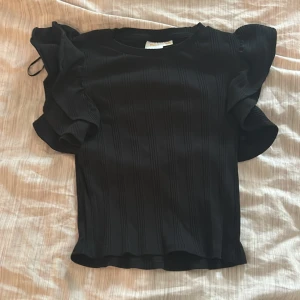 Svart topp med volangärmar från Gina Tricot - Säljer en svart topp från Gina Tricot med breda volangärmar som ger en cool och trendig look. Toppen har en lätt ribbad struktur och är figurnära i modellen. Perfekt för dig som vill sticka ut lite extra.