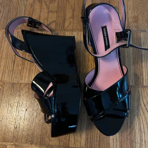 Svarta platåsandaler från Sonia Rykiel x H&M - Säljer ett par svarta glansiga platåsandaler från Sonia Rykiel x H&M. Skorna har öppen tå, bred klack och remmar som korsas framtill samt justerbar rem runt vristen. Insidan är ljusrosa för en snygg kontrast.