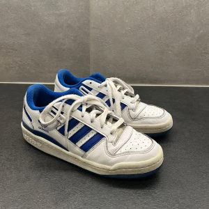 Blå Adidas sneakers Forum Low - Säljer ett par klassiska Adidas Forum Low sneakers i vitt med blå ränder och detaljer. Skorna har snörning och kardborreband upptill som kan tas bort (se bild). Storlek 41 1/3 men passar mig som är mellan 40-41. Har använts lite men är fortfarande i bra skick!💕Pris kan diskuteras!