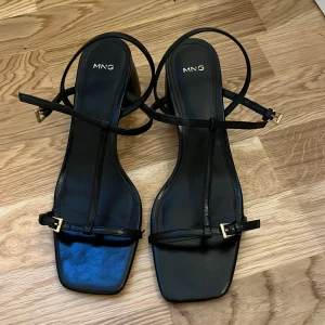Svarta sandaler med klack från MNG - Säljer ett par stilrena svarta sandaler från MNG med fyrkantig tå och smala remmar. Skorna har en medelhög blockklack och guldfärgade spännen vid ankeln. Använda en gång och är en ny modell. Även väldigt sköna som kan passa till en kvälls outfit!