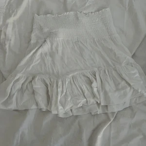 Vit volangkjol med smock - Supersöt vit kjol med bred smock i midjan och volang nertill. Kjolen är kort och har en luftig känsla, perfekt för varma dagar. Passar till många olika stilar och är lätt att matcha med olika toppar.