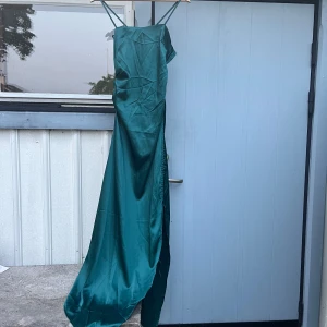 Grön långklänning med slits och smala axelband - Säljer en elegant grön långklänning i satinliknande material med smala axelband och slits på sidan. Klänningen faller fint längs kroppen. Perfekt för speciella tillfällen!