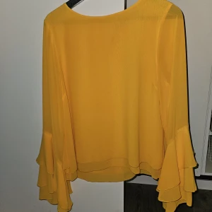 Gul blus med volangärmar från BikBok - Säljer en snygg gul blus från BikBok med långa volangärmar. Blusen har en lös passform och är tillverkad i ett lätt, flowy material som ger en luftig känsla. Perfekt för att liva upp garderoben med lite färg!