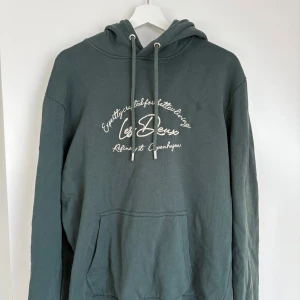 Mörkgrön hoodie från Les Deux - Säljer en mörkgrön hoodie från Les Deux med vit broderad text på bröstet. Tröjan har huva med dragsko och en stor magficka. Perfekt för en avslappnad stil. Använd typ 2 gånger.