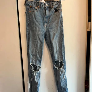 Blå  jeans med hål från Zara - Säljer ett par blå jeans från Zara med hål vid knäna. Byxorna har hög midja, raka ben och slits nertill. Klassisk femficksmodell i denim med råa kanter och trendig, avslappnad look.Tyvärr ett litet hål vid sidan men inget man tänker på om man har en tröja över, men därför sänks priser 