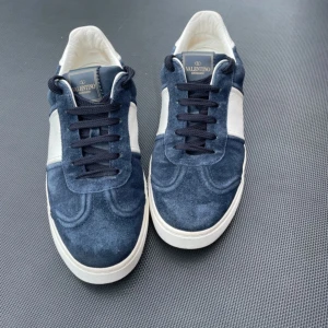 KÖP INTE NU! - Snygga sneakers från Valentino Garavani i blå mocka med vita detaljer och svarta skosnören. Skorna har rund tå, platt sula och coola nitar baktill på sulan. Perfekta för dig som gillar exklusiva och stilrena sneakers.