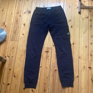 Stone Island byxor - Svarta mjukisbyxor från Stone Island Junior med klassisk logotyp-patch på benet. Byxorna har resår i midjan och vid bensluten samt en ficka på sidan med knapp. Perfekta för en avslappnad och stilren look. Har defekter, går att laga och tvätta bort.  
