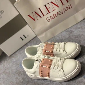 Vita sneakers Valentino Garavani Rockstud Untitled - Snygga vita sneakers från Valentino Garavani med ljusrosa band över vristen dekorerat med ikoniska pyramidformade nitar. Skorna har rund tå, platt sula och vita snören. Perfekta för dig som vill ha en lyxig och trendig look.