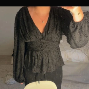 Svart blus - Säljer en svart blus med v-ringning och långärmade puffärmar. Blusen har markerad midja och volang nertill, samt ett strukturerat tyg med diskret mönster. Perfekt för dig som gillar unika detaljer och en feminin siluett.