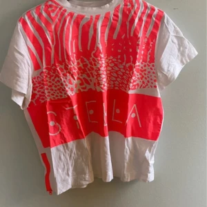 Vit t-shirt med rosa mönster från Stella McCartney - Vit t-shirt med stort neonrosa grafiskt mönster och texten 'STELLA' framtill. T-shirten har korta ärmar och en cool neonrosa dragkedja i sidan. Perfekt för dig som gillar färg och print. Nypris 500+ ❤️❤️