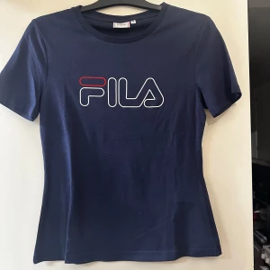 Mörkblå t-shirt från FILA - Säljer en mörkblå t-shirt från FILA med klassisk logga i vitt och rött framtill. T-shirten har rund halsringning och korta ärmar. Storlek M. Aldrig använd. 