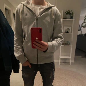 Grå Ralph lauren zip  - Grå hoodie med dragkedja från Ralph Lauren. Klassisk modell med huva och vita snören, samt broderad logga på bröstet. Perfekt för en avslappnad stil. Skick 9/10 använd ett fåtal gånger.