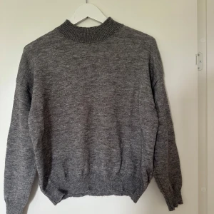 Grå stickad tröja med hög krage - Mysig grå stickad tröja med hög ribbad krage och långa ärmar. Tröjan har ribbade muddar vid ärmslut och nederkant, perfekt för kyligare dagar.