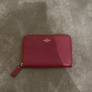 Röd plånbok från Valentino - Säljer en stilren röd plånbok från Valentino med guldfärgad dragkedja. Plånboken har flera kortfack och ett myntfack på insidan. Tillverkad i skinn med struktur och har diskret logga i guld framtill.