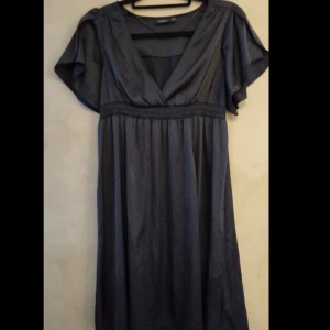 Svart silk klänning från Lindex - Svart klänning från Lindex med v-ringning och korta ärmar. Klänningen har ett markerat midjeband och ett mjukt, glansigt material som ger en elegant känsla. Perfekt för dig som gillar stilrena och klassiska plagg.