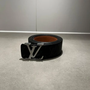 Svart LV bälte - Säljer ett svart bälte från Louis Vuitton med klassiskt monogrammönster och ett stort silverfärgat LV-spänne. Bältet är gjort i skinn och har en brun insida. Perfekt accessoar för att lyfta din outfit.