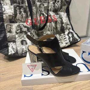 Svarta sandaler med klack från Guess - Säljer ett par svarta sandaler från Guess med öppen tå och breda remmar. Klacken är hög och dekorerad med ett coolt silvrigt nätmönster. Skorna har spänne vid ankeln och en modern, edgy look. Perfekta för dig som vill sticka ut.