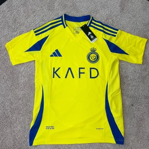 Al Nassr gul fotbollströja från Adidas - Säljer en gul Al Nassr fotbollströja från Adidas med blå detaljer och klubbmärke på bröstet. Tröjan har korta ärmar, blå ränder på axlarna och är tillverkad i lätt funktionsmaterial. Perfekt för fotbollsträning eller match.