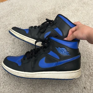Nike Air Jordan 1 Retro High OG svart/blå - Säljer ett par Nike Air Jordan 1 Retro High OG i svart och blått med klassisk Jordan-logga på sidan och blå detaljer. Skorna har hög modell, snörning och vit mellansula. Inköpta för 1400, använda men fortfarande väldigt fina skor