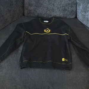 Svart sweatshirt från Dignitas med gula detaljer - Säljer en svart sweatshirt från Dignitas med gula sömmar och broderad logga framtill. Tröjan har en gul detalj vid nederkanten och en broderad emblem på bröstet. Perfekt för dig som gillar gaming och stilrena plagg.