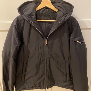 Prada Windbreaker Jacka - Säljer en svart windbreaker jacka från Prada. Jackan har en ficka med dragkedja på ärmen och klassisk Prada-logga. Perfekt för dig som vill ha en stilren och exklusiv look. Använd ett fåtal gånger i storlek L men passar de som är M också. 