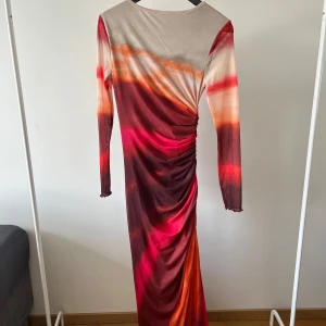 Färgstark långklänning med drapering - Säljer en långklänning med coola färgskiftningar i rött, orange, rosa och beige. Klänningen har lång ärm och snygg drapering längs sidan som ger en unik look. Perfekt för dig som vill sticka ut!