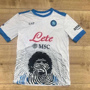 Napoli Maradona 10 fotbollströja - Säljer en vit Napoli fotbollströja med blå detaljer, Maradona 10 på ryggen och Serie A-märke på ärmen. Framsidan har klubbmärke, sponsorloggor och ett unikt fingeravtrycksmotiv. Perfekt för fans av Napoli och Maradona.