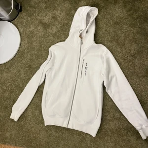 Vit hoodie från Sail Racing - Säljer en stilren vit hoodie från Sail Racing med dragkedja och huva. Jackan har en bröstficka med dragkedja och logga på framsidan.Jätte bra skick inga fläckar.Hör av er för fler frågor!!