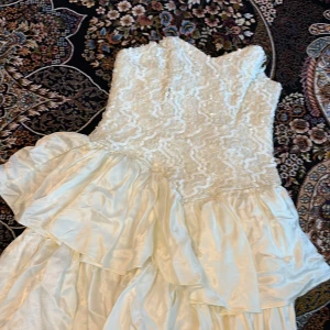 Vit långklänning med spets och volang från Scarlett Nite - Romantisk vit långklänning från Scarlett Nite med vacker spets upptill och paljettdetaljer. Klänningen har volanger i kjolen och en axelbandslös design som ger en elegant känsla. Perfekt för speciella tillfällen.