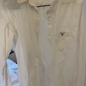 Vit skjorta från Gant - Klassisk vit skjorta från Gant med bröstficka och broderad logga. Skjortan har knappar framtill och lång ärm, perfekt för en clean och stilren look. Passar dig som gillar tidlös stil och enkelhet.