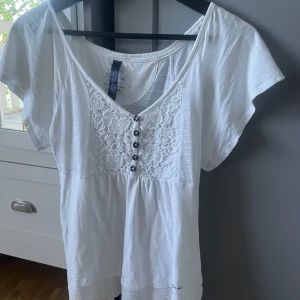 Vit blus med spets och knappar - Söt vit blus med korta ärmar och v-ringning. Framsidan har fina spetsdetaljer och dekorativa knappar. Perfekt till sommaren och enkel att matcha med jeans eller kjol.