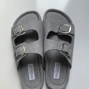 Grå sandaler med dubbla spännen - Säljer ett par grå sandaler från Primark med två breda remmar och silvriga spännen. Ovandelen är i filtliknande material och sulan är räfflad för extra grepp. Perfekta för sommaren och riktigt sköna att glida runt i.