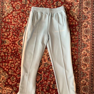 Ljusblå mjukisbyxor från Palm Angels - Säljer ett par ljusblå mjukisbyxor från Palm Angels med vita sidoränder och broderad logga på ena benet. Byxorna har elastisk midja och raka ben, perfekta för en avslappnad streetwear-look. Längd 104 cm.