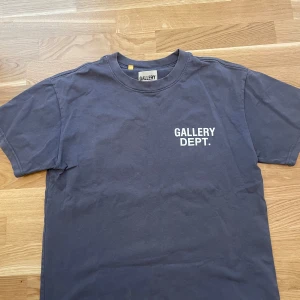 Mörkgrå t-shirt från Gallery Dept. - Säljer en mörkgrå t-shirt från Gallery Dept. med vit logga tryckt på bröstet. Klassisk rund halsringning och normal passform. Tillverkad i 100% bomull, perfekt för en avslappnad stil.