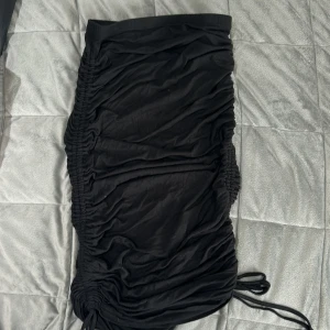Svart tight kjol med snörning - Säljer en svart tight kjol med snörning på sidorna som ger en snygg rynkad effekt. Kjolen är kort och har en elastisk midja. Perfekt för dig som vill ha en trendig och bekväm look.