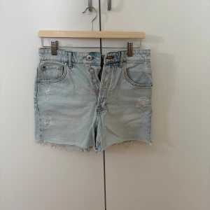Ljusblå jeansshorts med fransar - Säljer ett par ljusblå jeansshorts med slitna detaljer. Shortsen har fransig kant nedtill, klassiska fem fickor och knappar framtill. Perfekta för varma dagar och en avslappnad stil.