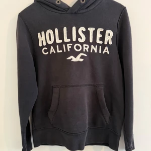 Svart hoodie från Hollister - Svart hoodie från Hollister med vit broderad logga och text på bröstet. Klassisk känguruficka framtill och huva med dragsko. Perfekt för en avslappnad och stilren look. Jättefin tröja. 