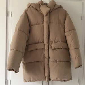 Beige pufferjacka från Zara - Säljer en beige pufferjacka från Zara i storlek 164. Jackan har huva, dragkedja och stora fickor framtill. Perfekt för kyligare dagar och har en klassisk, enkel design som passar till det mesta. Aldrig använd.