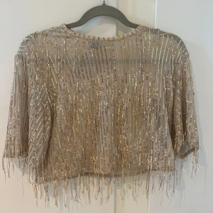 Glittrig croppad topp med paljetter - Säljer en beige croppad topp med glittriga paljetter i guld och silver. Toppen har trekvartsärm och fransar längst ner för extra effekt. Perfekt för dig som vill sticka ut på festen! OBS tröjan är genomskinlig så ett linne eller nån slags topp/bh behövs under