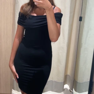 Svart offshoulder klänning - Säljer en svart, tight klänning med offshoulder-design. Klänningen har en smickrande passform och är perfekt för dig som gillar en stilren och enkel look. Materialet känns mjukt och följsamt mot kroppen.
