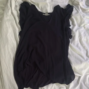 Mörkblå volangklänning med v-ringning - Säljer en mörkblå tröja i storlek M med v-ringning och volangdetaljer vid ärmarna. Klänningen har en luftig och lätt känsla, perfekt för varmare dagar. Enkel och stilren design som passar till många tillfällen.