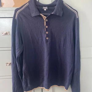 Mörkblå pikétröja från D&G - Snygg mörkblå pikétröja från D&G med långa ärmar och krage. Tröjan har knappar framtill och diskreta beige detaljer längs krage och axlar. Perfekt för en stilren look.