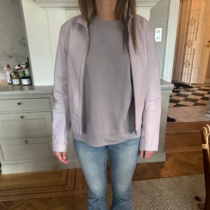 Ljuslila skinnjacka - Säljer en ljuslila skinnjacka med minimalistisk design. Jackan har lång ärm, ståkrage och dragkedja framtill. Passformen är normal och jackan har diskreta spänndetaljer i sidorna. Perfekt för dig som vill ha en stilren look.❤️❤️❤️