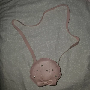 Ljusrosa axelväska med pärlor och rosett - Supergullig ljusrosa axelväska i form av en snäcka, dekorerad med små pärlor och en söt rosett framtill. Väskan har ett långt axelband och är perfekt för att bära småsaker. Baksidan är slät och enkel.