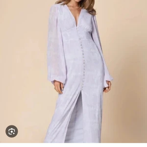 Lila Adoore klänning - Säljer en elegant ljuslila långklänning med v-ringning och långa ärmar. Klänningen har dekorativa knappar framtill och en slits som ger en snygg detalj. Perfekt för speciella tillfällen!