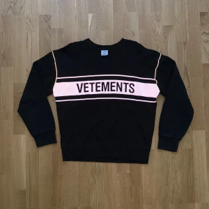 Svart sweatshirt från Vetements size M-L reflection  - Säljer en svart sweatshirt från Vetements med vit rand och stor logga över bröstet. Tröjan har rund halsringning och långa ärmar. Perfekt för en avslappnad och trendig stil.