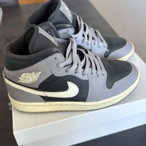 Nike Air Jordan 1 Mid i grå och svart - Säljer ett par Nike Air Jordan 1 Mid i grå och svart med vita detaljer. Skorna har klassisk snörning, rund tå och den ikoniska Air Jordan-loggan på sidan. Perfekta för dig som gillar streetwear och sneakers med stil.